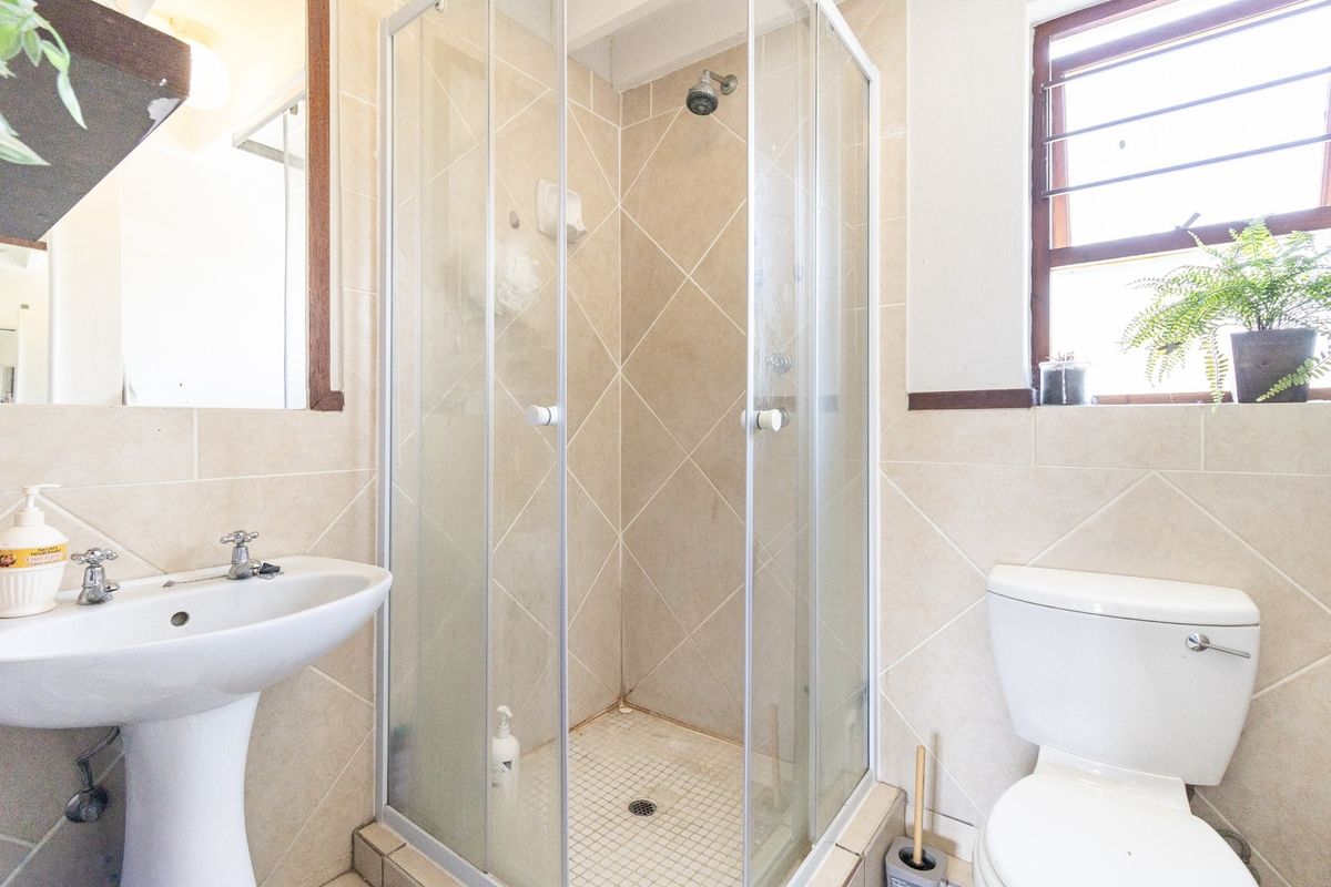 A functional bathroom featuring a pedestal washbasin, a corner shower enclosure with glass doors, and a standard toilet, finished with neutral wall and floor tiles.