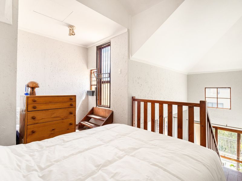 A loft-style bedroom featuring a timber balustrade, vaulted ceilings, and textured wall finishes, overlooking the lower level of the apartment.
