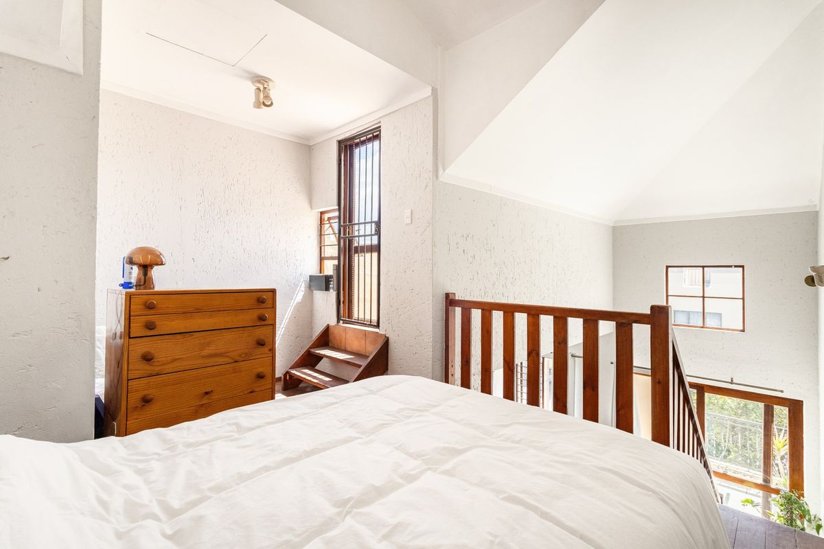 A loft-style bedroom featuring a timber balustrade, vaulted ceilings, and textured wall finishes, overlooking the lower level of the apartment.