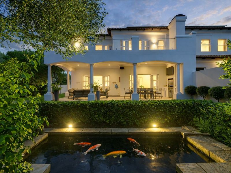 Rear view of a two-story residence featuring a covered patio with built-in braai, overlooking a landscaped garden with a stone-edged koi pond.