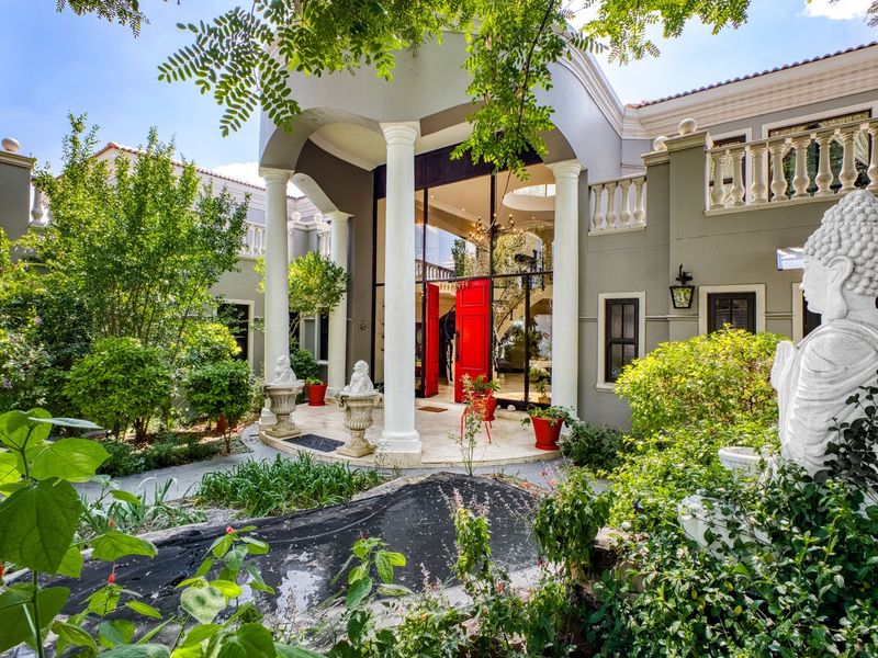 Grand front entrance featuring a covered portico with classical columns, double-height glass doors, and a stone-paved entryway surrounded by lush garden landscaping.