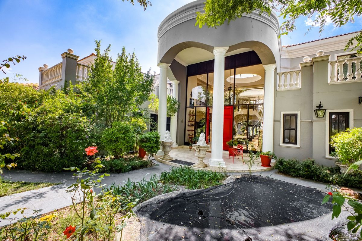 A grand rear exterior view featuring a covered patio with classical columns, stone paving, and a well-maintained garden setting.