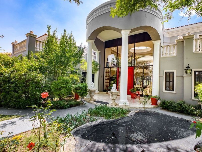 A grand rear exterior view featuring a covered patio with classical columns, stone paving, and a well-maintained garden setting.
