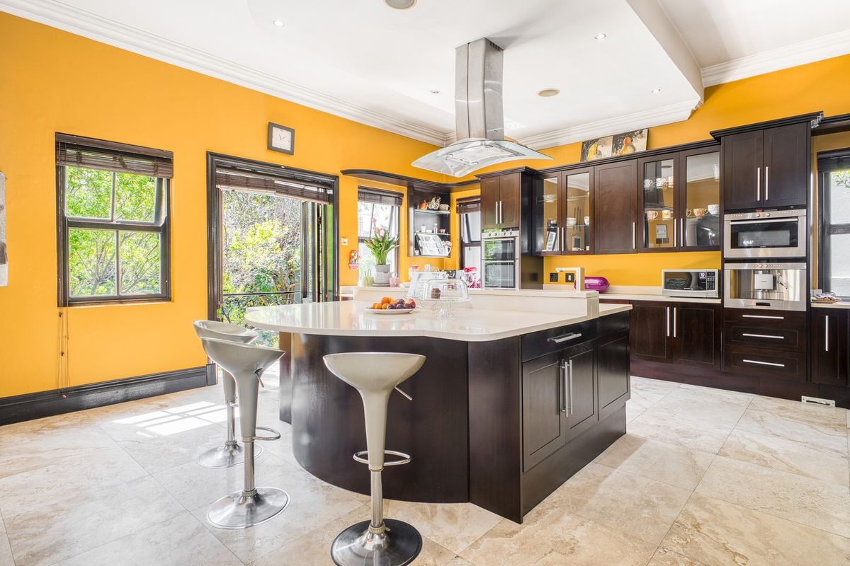 Spacious kitchen featuring dark wood cabinetry, a large central island with stone countertops, stainless steel appliances, and bright yellow walls with direct garden access.