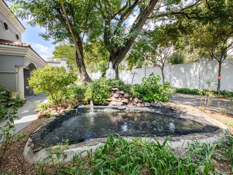 A tranquil garden area featuring a central pond with a water fountain, mature trees, and a paved walkway alongside the property exterior.
