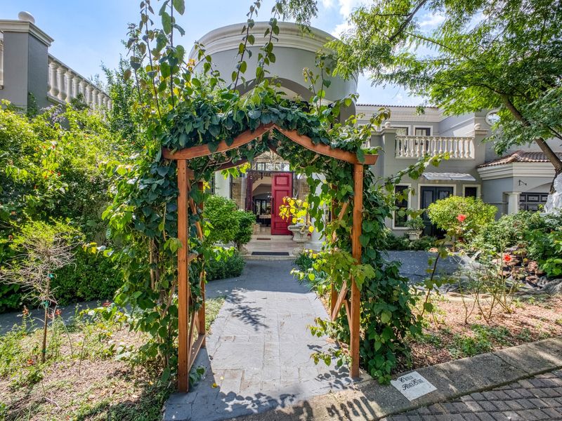 A charming front entrance featuring a stone paved walkway, a decorative wooden garden arch, and a classic double-storey facade with elegant balustrades.