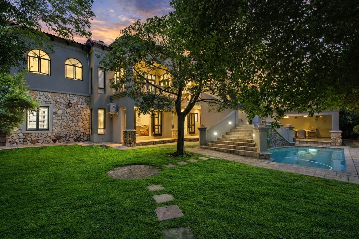 A beautifully landscaped rear garden featuring a swimming pool, stone-clad facade, covered patio, and an elegant outdoor staircase leading to the upper level.