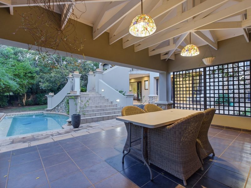 Covered outdoor patio featuring tiled flooring, exposed timber rafters, and pendant lighting, overlooking a swimming pool and stone staircase in the garden.