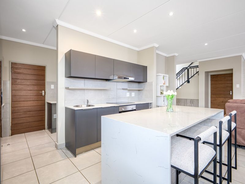 Modern open-plan kitchen featuring sleek dark cabinetry, a stone-topped island, integrated appliances, and tiled flooring, seamlessly connecting to the living area and staircase.