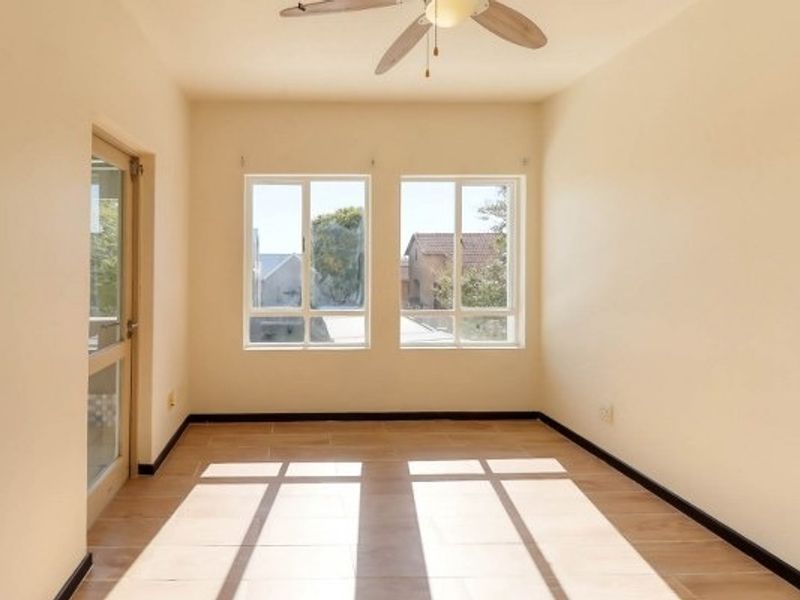 A bright, empty bedroom featuring tiled flooring, a ceiling fan, and natural light through double windows with a glass panel door.