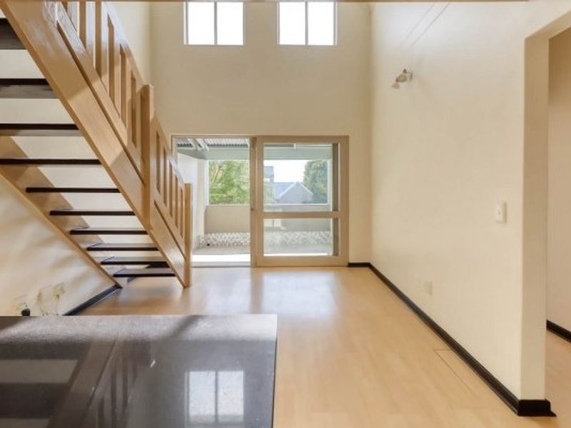 Spacious double-volume living area featuring light laminate flooring, a wooden staircase, and sliding glass doors leading to an outdoor balcony.
