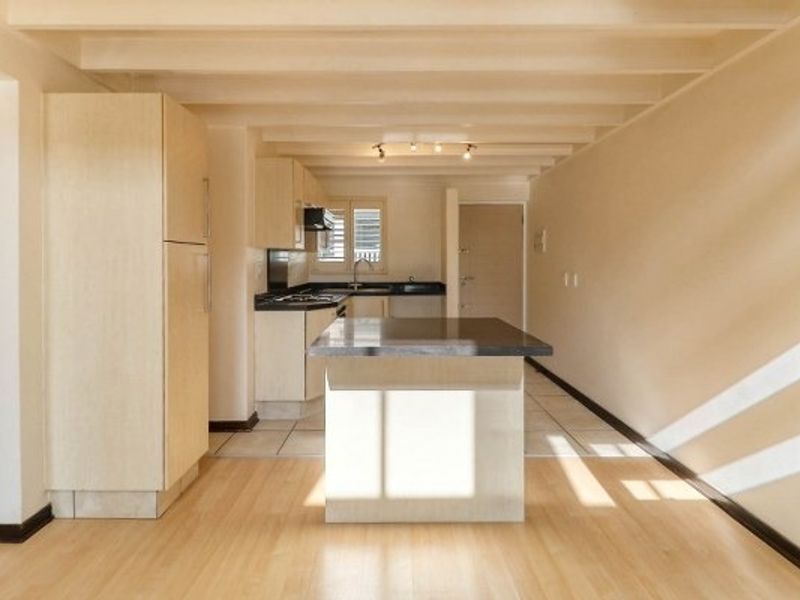 An open-plan kitchen area featuring light wood cabinetry, a central island, laminate flooring, and exposed ceiling beams in a bright, vacant space.