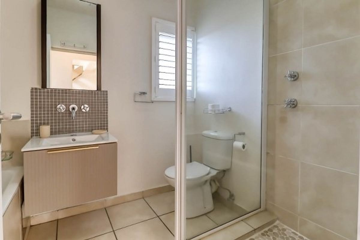 A functional bathroom featuring a vanity with mosaic splashback, a glass-enclosed shower, and a toilet, finished with neutral floor tiles and a shutter window.