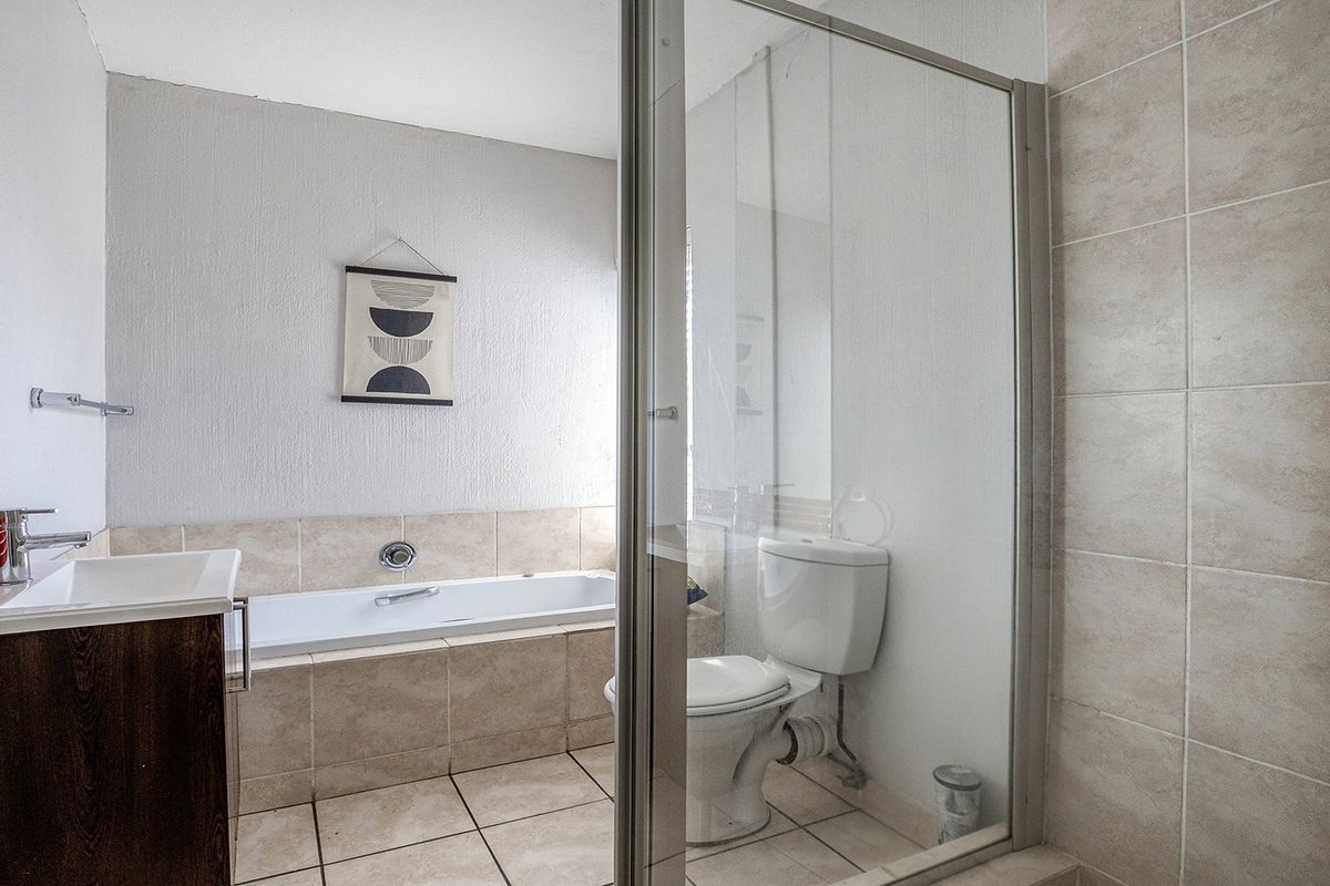 A functional bathroom featuring a built-in bathtub, vanity with basin, toilet, and a separate glass-enclosed shower area with neutral wall and floor tiling.