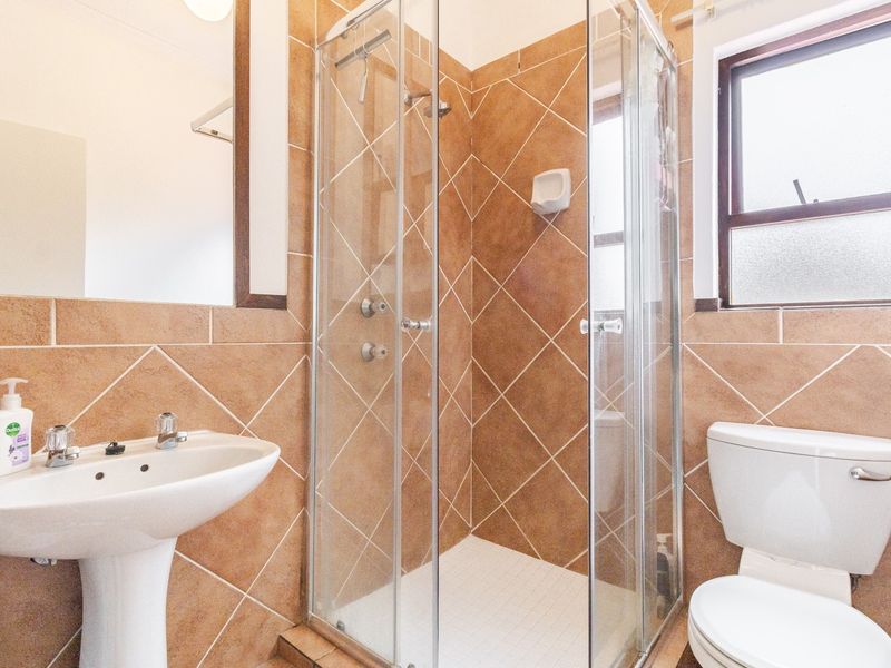 A functional bathroom featuring a pedestal basin, a glass-enclosed shower, and a toilet, finished with neutral-toned wall and floor tiles.