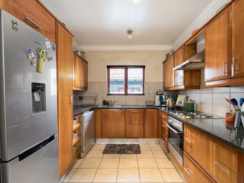 A functional galley-style kitchen featuring warm wooden cabinetry, granite countertops, integrated stainless steel appliances, and neutral tiled flooring.