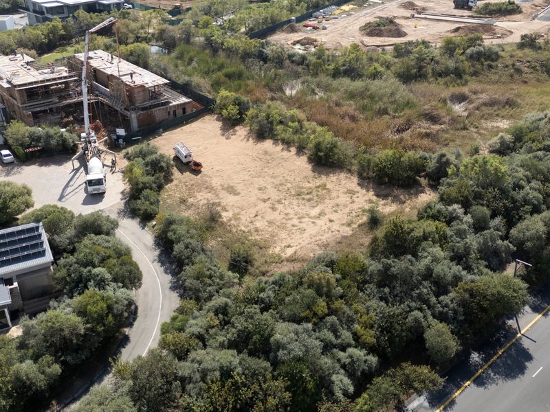 Aerial view of a vacant residential plot surrounded by established vegetation and adjacent to a paved access road in a developing estate.