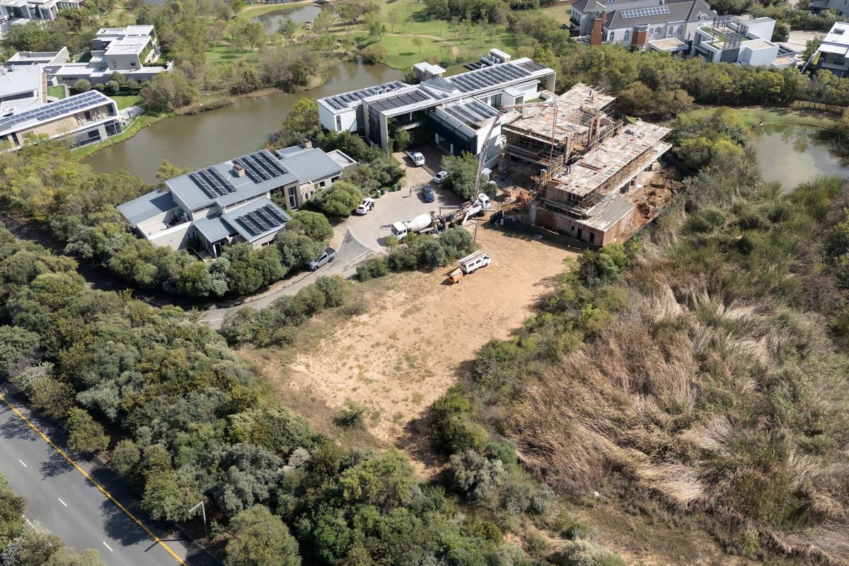 Aerial view of a vacant residential plot situated within a developed estate, featuring surrounding natural landscape and nearby ongoing construction.