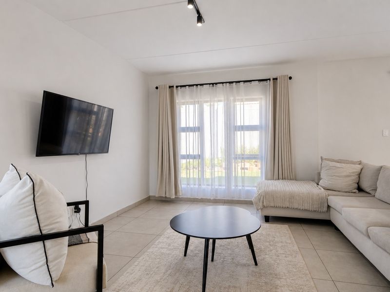 A bright and modern living room featuring neutral tiled flooring, minimalist track lighting, and large windows allowing for ample natural light.
