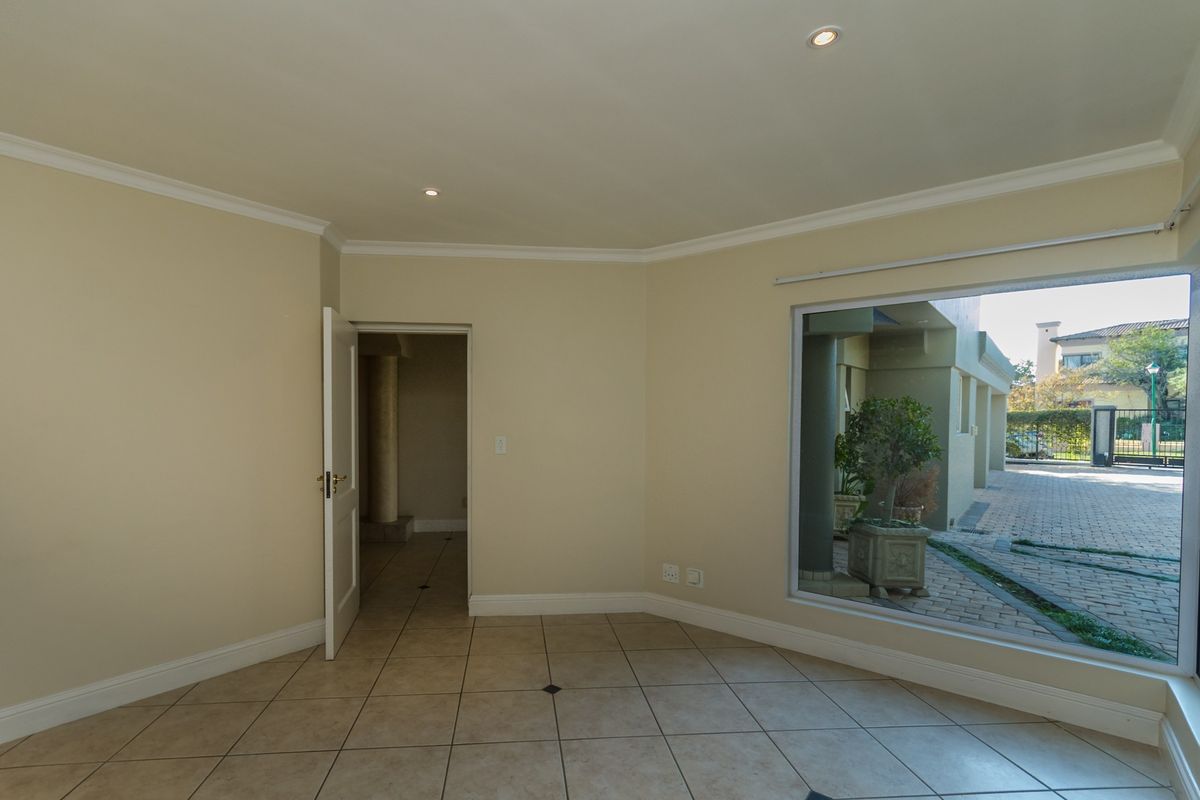 A bright, empty living area featuring neutral tiled flooring, recessed downlighting, and a large window providing a view of the exterior paved area.
