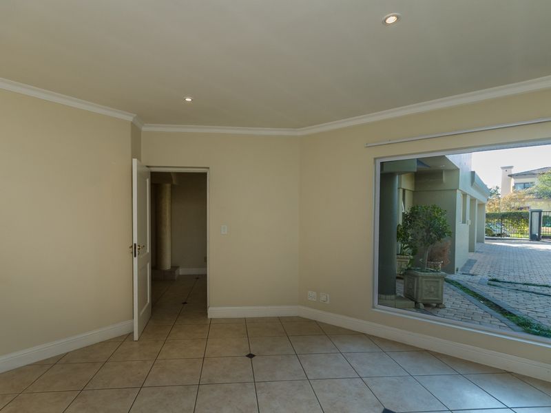 A bright, empty living area featuring neutral tiled flooring, recessed downlighting, and a large window providing a view of the exterior paved area.