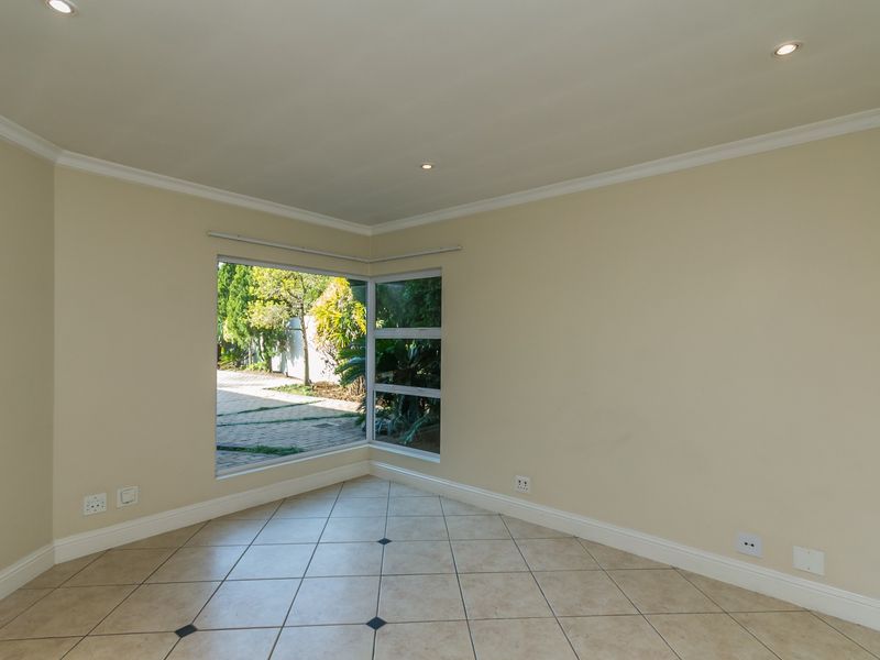 A bright, empty living area featuring neutral walls, tiled flooring, and a large corner window providing natural light and garden views.