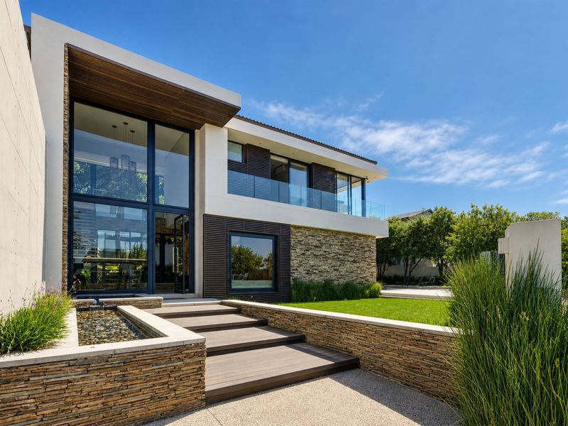 Modern architectural front facade featuring stone cladding, timber decking, a tranquil water feature, and expansive double-volume glass windows.