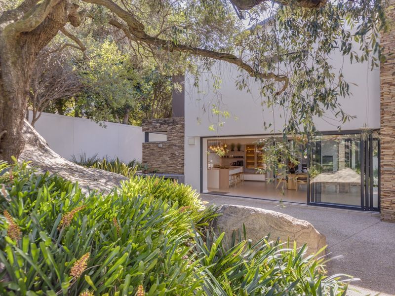A modern rear exterior view featuring stacked stone accents, large sliding glass doors opening to a paved patio, and a lush, landscaped garden setting.