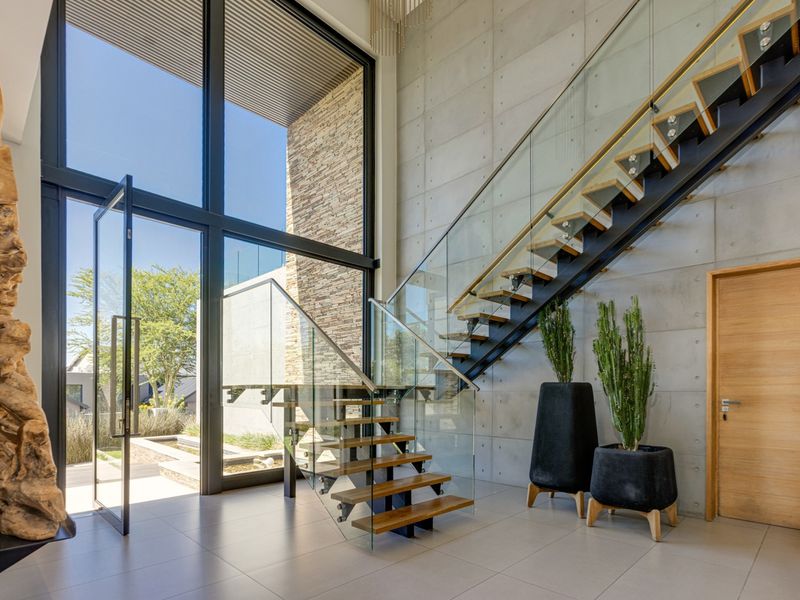 A grand double-volume entrance hall featuring a modern floating staircase with glass balustrades, concrete feature walls, and expansive floor-to-ceiling windows.