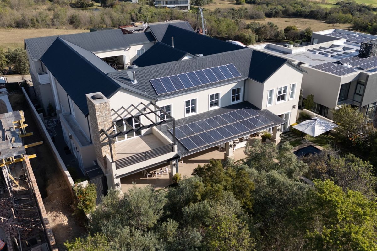 Aerial view of a modern residential property featuring a metal roof with integrated solar panels, a stone chimney, and a spacious covered patio area.