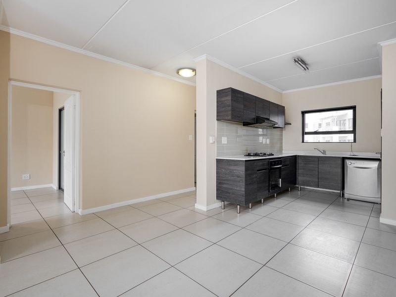 A clean, modern kitchen featuring dark wood-look cabinetry, a gas hob, built-in oven, and neutral tiled flooring in an open-plan layout.