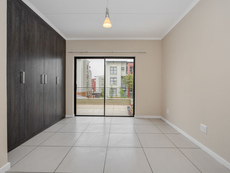 A bright, empty bedroom featuring dark wood built-in cupboards, neutral tiled flooring, and a sliding glass door leading to a private balcony.
