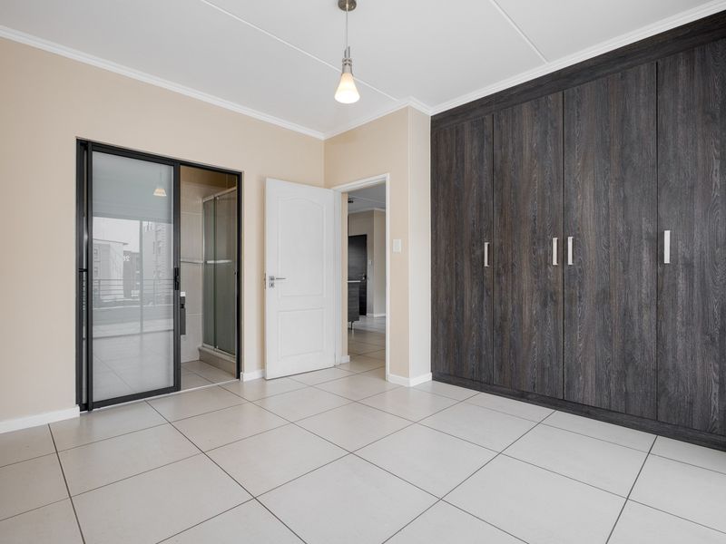 A spacious, empty bedroom featuring modern dark wood built-in cupboards, neutral tiled flooring, and direct access to an en-suite bathroom.
