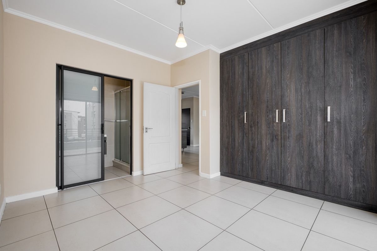 A spacious, empty bedroom featuring modern dark wood built-in cupboards, neutral tiled flooring, and direct access to an en-suite bathroom.