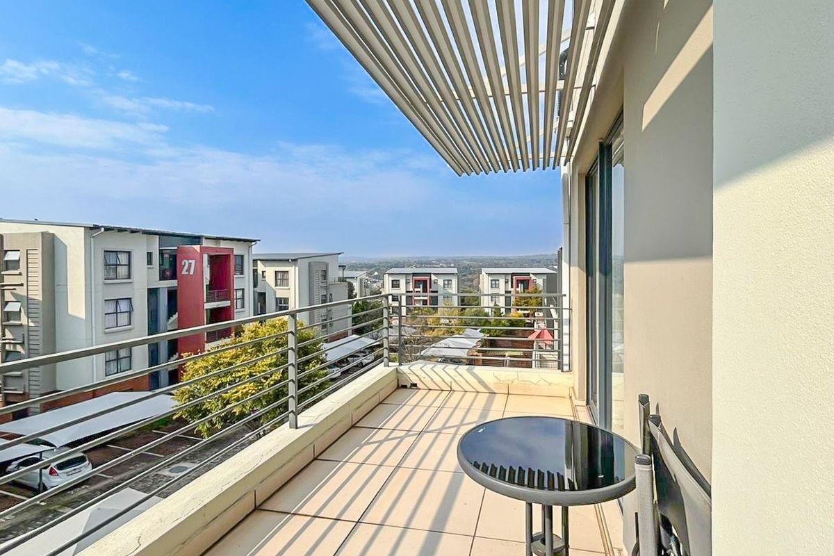 A modern apartment balcony featuring tiled flooring, a protective slatted pergola, and metal railings, offering an elevated view of the surrounding residential complex.