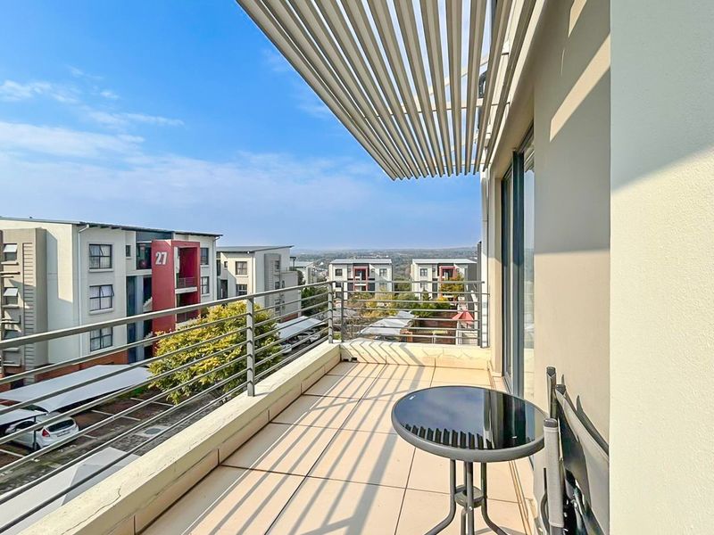 A modern apartment balcony featuring tiled flooring, a protective slatted pergola, and metal railings, offering an elevated view of the surrounding residential complex.