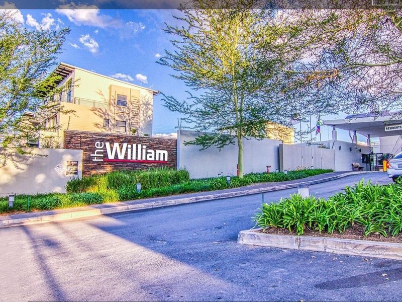 The entrance to The William apartment complex, featuring a modern security gate, landscaped gardens, and a paved driveway.