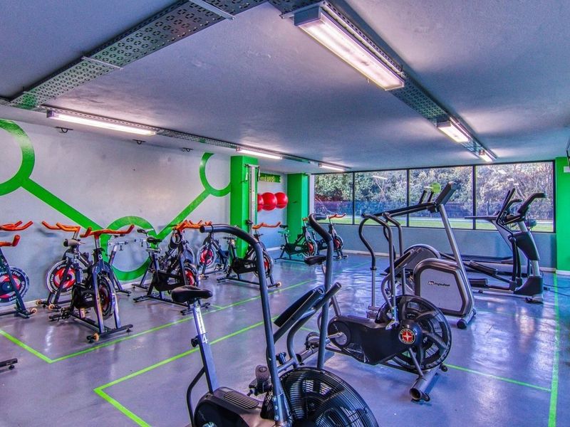 A functional apartment complex gym featuring painted concrete floors, exposed industrial cable trays, fluorescent lighting, and large windows overlooking the exterior.