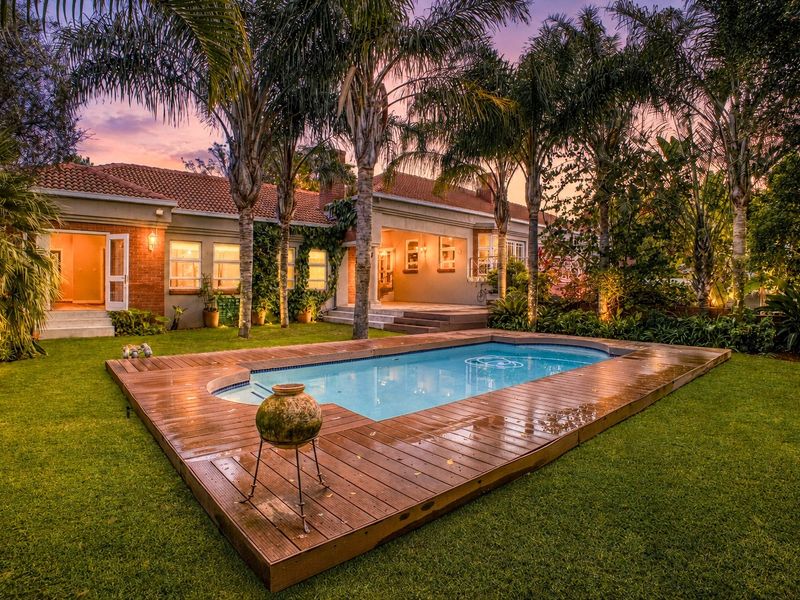 A beautifully landscaped backyard featuring a swimming pool with a timber deck, surrounded by mature palm trees and a well-maintained garden at dusk.
