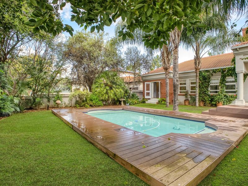 A well-maintained backyard featuring a swimming pool with timber decking, surrounded by a lush garden and the rear brick facade of the house.