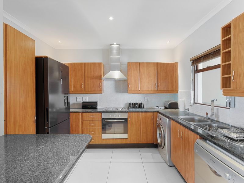 A functional kitchen featuring wood-finish cabinetry, granite countertops, built-in oven with gas hob, stainless steel extractor fan, and tiled flooring.