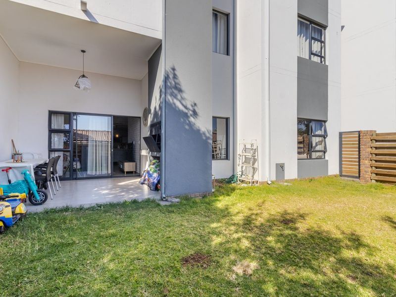 Rear view of the apartment featuring a covered patio with a built-in braai, tiled flooring, and a private garden area with timber fencing.