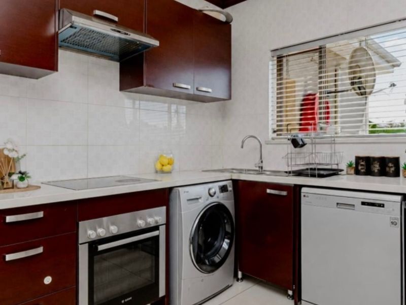 Compact kitchen featuring dark wood cabinetry, integrated oven and hob, stainless steel sink, and tiled finishes with space for laundry and dishwasher appliances.