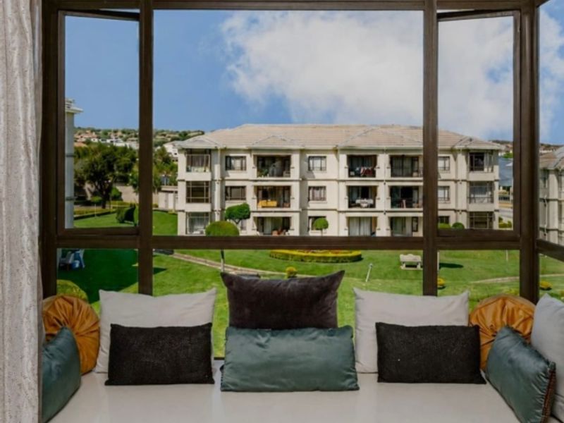 A bright enclosed balcony featuring a built-in window seat with storage cabinetry, offering pleasant views of the surrounding apartment complex and landscaped grounds.