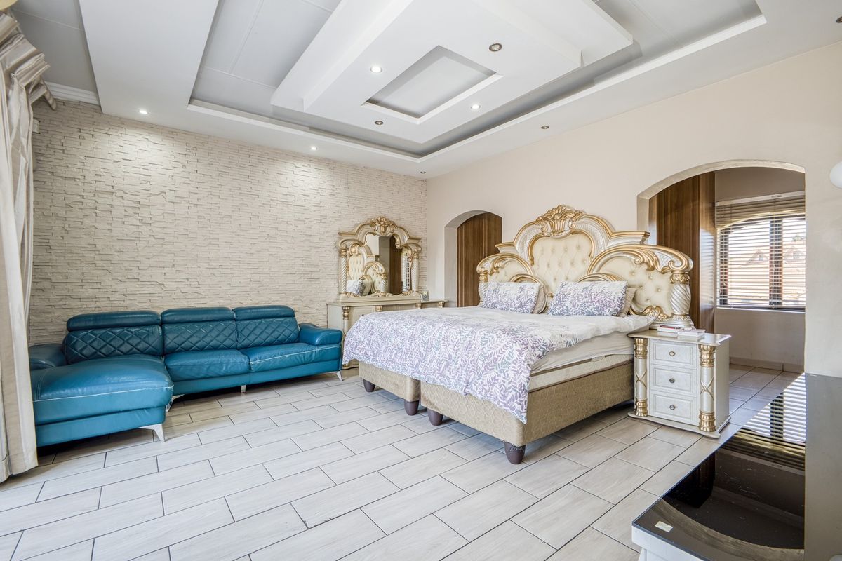 Spacious bedroom featuring a decorative recessed ceiling, stone-clad feature wall, and neutral tiled flooring with an integrated archway leading to an adjacent area.