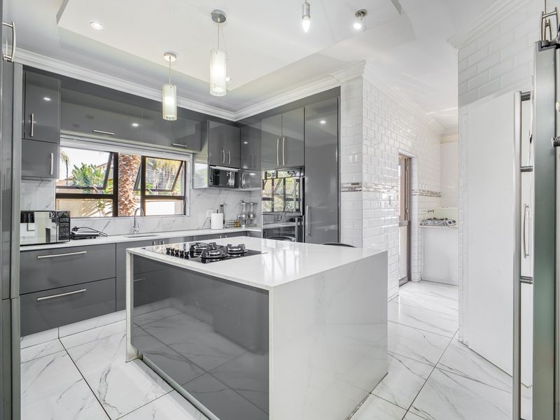 Modern kitchen featuring a central island with gas cooktop, sleek grey cabinetry, marble-look flooring, and subway tile backsplash, leading to a separate scullery area.