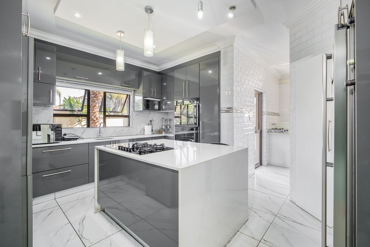 Modern kitchen featuring a central island with gas cooktop, sleek grey cabinetry, marble-look flooring, and subway tile backsplash, leading to a separate scullery area.