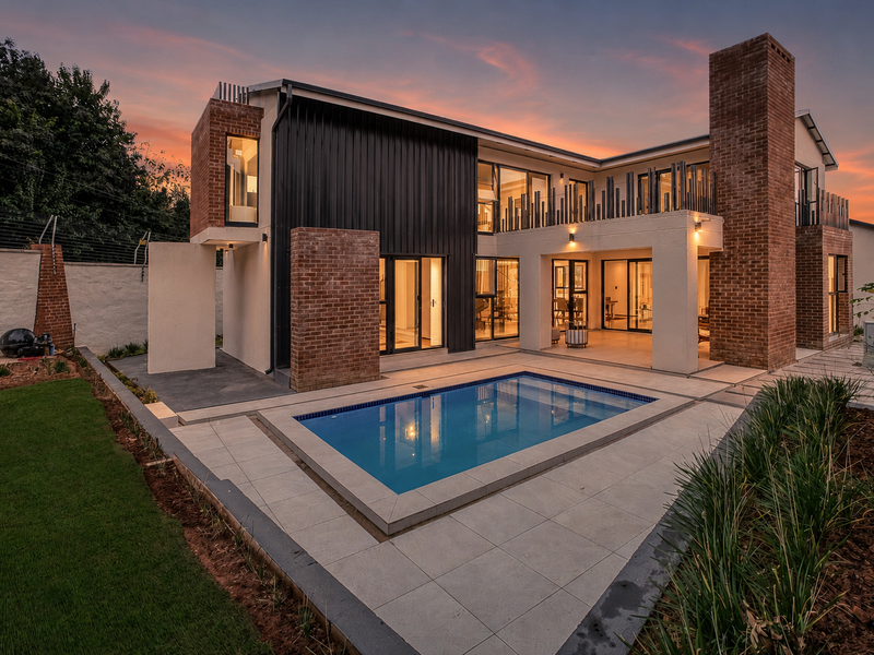 Modern double-story residence featuring a rear swimming pool, covered patio, balcony, and a striking combination of brick and metal cladding at sunset.
