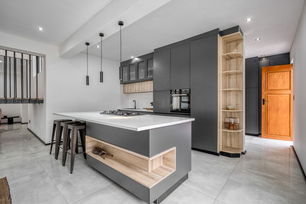 Modern kitchen featuring a central island with gas hob, sleek dark cabinetry, integrated oven, and light wood shelving accents on polished tiled floors.