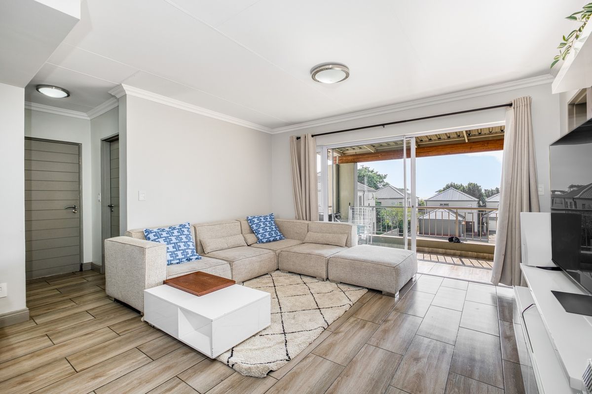 A bright and well-maintained living room featuring wood-look tiled flooring, modern ceiling lighting, and a large sliding door leading to an outdoor balcony.